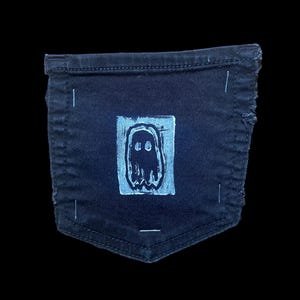 May include: Dark blue denim pocket featuring a light blue ghost design. The ghost is printed on a light blue square. The pocket is from a pair of jeans.