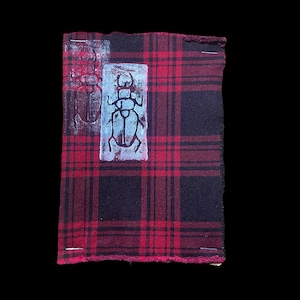 May include: A section of red and black tartan fabric featuring two insect prints. One print is faded red, the other is a blue insect print on a white rectangle. The fabric is held together with silver pins.