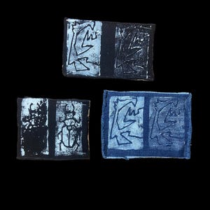May include: Three abstract art pieces against a black backdrop. Each is a rectangular patch with a dark background and a white, stylised design. The designs appear to be linocut prints, with one featuring a scarab beetle.