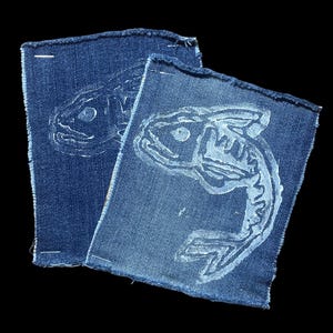 Peut inclure: Deux écussons en denim bleu avec un motif de poisson blanc. Le poisson est stylisé avec des os et des nageoires visibles. Les bords des écussons sont effilochés, sur fond noir.