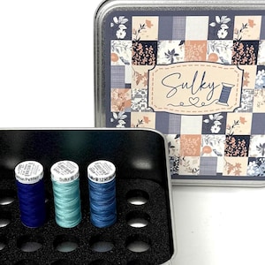 Puede incluir: Una caja de almacenamiento de hilo de coser de metal plateado con tres carretes de hilo azul. La caja tiene un inserto de espuma negra. La tapa presenta un diseño de patchwork con la palabra "Sulky" y una bobina de hilo.