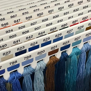 Op de afbeelding: Een close-up van een verzameling DMC borduurgaren spoelen, georganiseerd op kleur nummer. De spoelen zijn gerangschikt in rijen, met de nummers zichtbaar op elke spoel. De kleuren variëren van lichtblauw tot bruin.