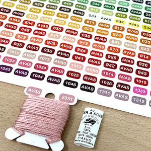 Puede incluir: Primer plano de un hilo de bordar rosa enrollado en una bobina de plástico blanca. La bobina está etiquetada con "2931". Una pequeña etiqueta blanca con texto negro dice "Au fil a soie d'Alger 5m 100% soie - seide seta - seda Paris Made in France".