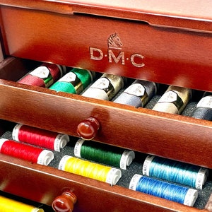 May include: A wooden box with three drawers filled with spools of DMC embroidery floss in various colours. The box has a wooden handle and a brass nameplate that reads "D.M.C."