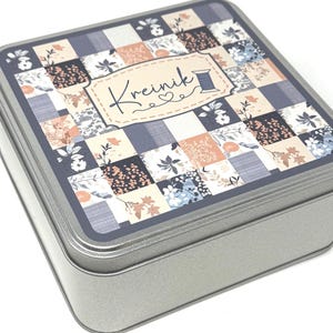 May include: A square metal tin with a patchwork design lid. The lid features a pattern of squares in shades of blue, peach, and white with floral accents. The text "Kreinik" is displayed on the lid, along with a spool of thread graphic.