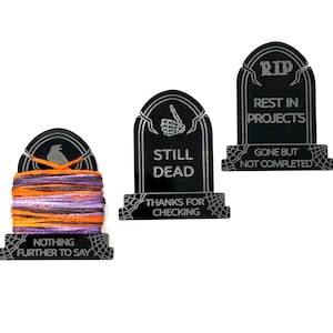 May include: Three black tombstone-shaped decorations with white text and spiderweb accents. One tombstone has a spool of orange, purple, and white thread. The text reads "Nothing Further to Say", "Still Dead", and "Rest in Projects".