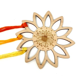 May include: A wooden sunflower-shaped embroidery floss organizer with 12 slots. The slots are numbered 1 through 12. Three strands of floss are threaded through the slots.