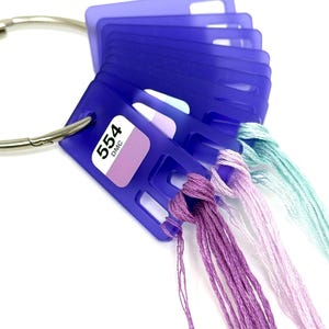 Könnte beinhalten: Nahaufnahme eines violetten Plastik-Stickgarn-Organizers mit einem Metallring. Der Organizer hat ein Etikett mit der Aufschrift "554 DMC". Der Organizer enthält mehrere Fäden in verschiedenen Schattierungen von Lila, Rosa und Blau.