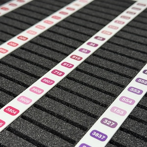 May include: Close-up of a black foam surface with white strips featuring numbers in various colors. The numbers are in shades of pink, purple, and red. The strips are evenly spaced across the surface.