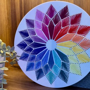 May include: A circular wall decoration with a vibrant, geometric pattern. The design is made with threads in a rainbow of colours, emanating from the centre. Gold-coloured details are also visible.