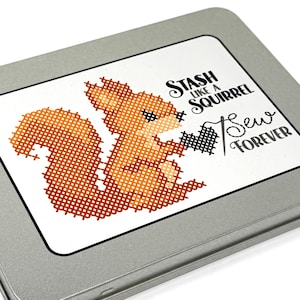 Op de afbeelding: Een zilveren metalen doos met een wit etiket met een kruissteekontwerp van een eekhoorn met een naald en draad. De tekst luidt "Stash Like a Squirrel & Sew Forever."