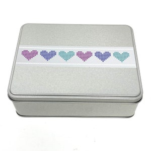 Może przedstawiać: Srebrna metalowa puszka z białym paskiem, na którym znajduje się rząd kolorowych haftowanych serc. Serca są w odcieniach różowego, fioletowego, niebieskiego i zielonego.