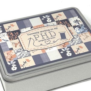 Może przedstawiać: Prostokątne metalowe pudełko z pokrywą w patchworkowy wzór. Pokrywa zawiera mieszankę wzorów kwiatowych i geometrycznych w odcieniach niebieskiego, brzoskwiniowego i kremowego. Tekst na pokrywie brzmi "Currently working on my PHD (projects half done)" z ilustracją szpulki nici.