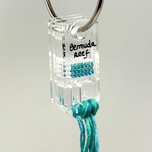 Könnte beinhalten: Transparenter Acryl-Schlüsselanhänger mit einem silbernen Ring. Der Schlüsselanhänger hat eine rechteckige Form mit den Worten "Bermuda Reef" darauf geschrieben. Er hat ein türkis- und teal-farbenes Design und eine Quaste aus teal-farbenen Fäden.