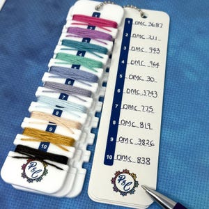 Puede incluir: Una tarjeta blanca de colores de hilo con diez muestras de color, cada una con un número DMC correspondiente. Los colores varían del rosa al negro. Un bolígrafo descansa cerca de la tarjeta. La tarjeta tiene un logotipo con las letras P&C.