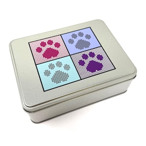 May include: A silver metal tin with a paw print design. The lid features four squares, each with a different coloured paw print: pink, blue, purple, and light blue. The paw prints are made of a cross-stitch pattern.