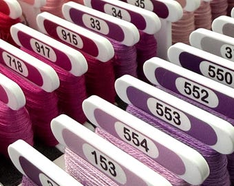May include: A collection of embroidery floss skeins wrapped around white plastic bobbins, organized in rows. Each bobbin features a colored label with a printed numerical identifier, showcasing a gradient of pink, purple, and lavender thread colors.
