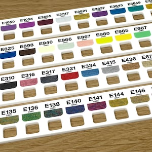 Peut inclure: Gros plan sur un organisateur en plastique blanc avec des rangées de petits carrés contenant du fil à broder de différentes couleurs. Chaque carré est étiqueté avec un numéro DMC et un code couleur.