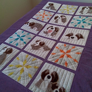 May include: A handmade quilt featuring squares with dog appliqués and starburst designs. The dogs are various breeds, with brown and white fur. The quilt has a purple border and is on a purple surface.