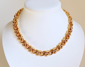 Könnte beinhalten: Eine goldfarbene Halskette mit herzförmigem Kettendesign. Die Halskette wird auf einer weißen Mannequin-Büste präsentiert. Die Glieder sind eng miteinander verbunden und bilden eine durchgehende Kette. Die Halskette hat eine polierte Oberfläche.