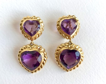 May include: A pair of gold-toned earrings featuring a heart-shaped amethyst gemstone. The earrings have a double-heart design, with a smaller heart above a larger heart. The gold setting has a rope-like border.