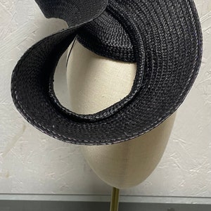 May include: A black woven hat with a unique, twisted design. The hat is displayed on a gold stand.
