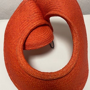 May include: A bright orange woven sun hat with a large brim and a unique, folded design. The hat is made of a straw-like material and has a wide, open space in the centre.