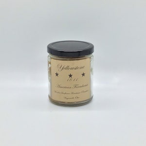 Peut inclure: Une bougie en verre dans un pot avec un couvercle noir. L'étiquette est brune avec du texte blanc qui dit "Yellowstone 1841 American Farmhouse Wooden Sunflower Farmhouse Scent Waynesville Ohio".