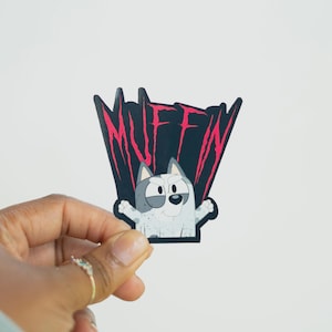 May include: A sticker featuring a cartoon dog character with grey and white fur, arms outstretched. The sticker has the word "MUFFIN" in pink, stylized text against a black background.