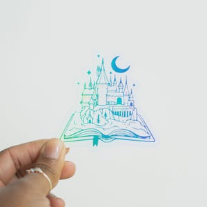 May include: A sticker featuring a castle design with a crescent moon and stars. The castle is set on an open book, with a gradient of blue and green colors. The sticker is held by a hand.