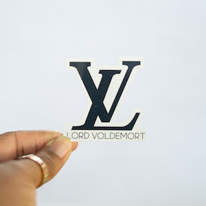 Puede incluir: Una pegatina en blanco y negro con un logotipo estilizado y el texto "LORD VOLDEMORT" debajo. El logotipo parodia el diseño de una marca conocida. La pegatina se sostiene sobre un fondo blanco.