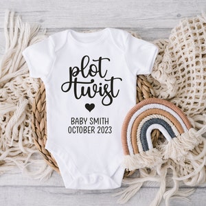 May include: A white baby grow with black text that reads "plot twist" and a small black heart. Below the text is "Baby Smith October 2023".