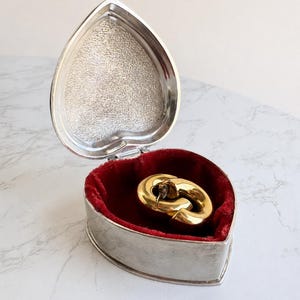 Puede incluir: Un par de pendientes de aro dorados pulidos dentro de una caja de metal plateado con forma de corazón. El interior de la caja está forrado con terciopelo rojo intenso y la tapa presenta un acabado plateado texturizado y brillante.