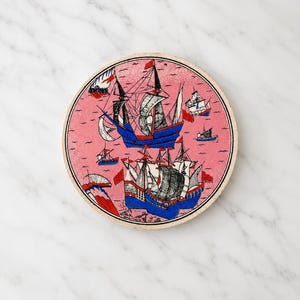 May include: A circular ceramic coaster featuring a pink background with a detailed illustration of multiple sailing ships. The ships are rendered in blue, black, and white with red accents on the flags and sails, depicting a nautical scene.