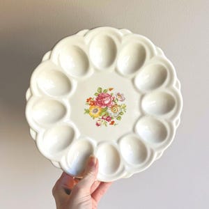 May include: A round, off-white ceramic egg serving platter with a scalloped edge and twelve oval indentations. The center of the platter features a colorful floral decal with pink, yellow, and white flowers and green leaves.