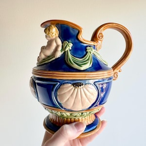 May include: A ceramic pitcher featuring a deep blue glaze with raised relief details in tan and green. The design includes a sculpted cherub figure near the rim, draped swags, and large shell motifs around the body, finished with a curved handle.