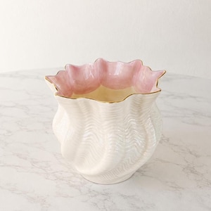 May include: A ceramic vase with a ruffled, scalloped rim featuring a soft pink interior and a cream-colored exterior. The body of the vase has a textured, wavy, ribbed pattern throughout, and the top edge is finished with a thin gold-toned trim.