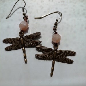 Pode incluir: Um par de brincos de libélula. Cada brinco apresenta um pingente de libélula de cor bronze suspensa sob uma conta de pedra preciosa cor de pêssego. Os brincos têm um fecho de gancho simples.