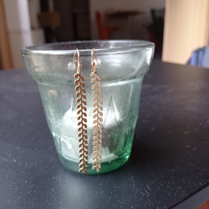 May include: Gold-colored leaf-shaped earrings hanging in front of a green glass. The earrings have a long, slender design with a series of leaf-like cutouts. The earrings are suspended in front of a green glass.