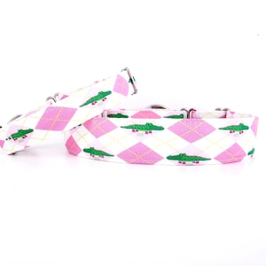 May include: Two dog collars with a pink argyle pattern and green alligator illustrations. The collars have silver metal hardware and are set against a white background. The argyle pattern is composed of pink diamonds and white lines.