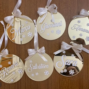 Può includere: Sei palline di Natale dorate con nomi in corsivo bianco e dettagli a fiocco di neve. Ogni ornamento ha un nastro e un fiocco bianchi. I nomi includono Marco, Giovanni, Annamaria, Rosaria, Salvatore e Sara.