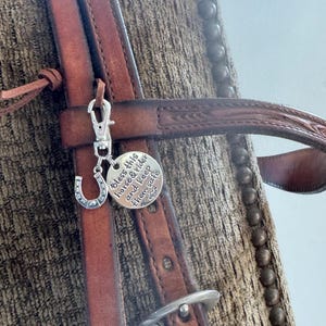 Op de afbeelding: Close-up van een bruine leren paardenhoofdstel met zilveren details en een bedel. De bedel heeft een hoefijzer en een tag met de tekst "Bless this horse & rider and keep them safe".