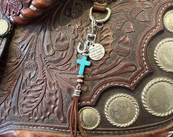 Op de afbeelding: Een bruin leren zadel met ingewikkelde bewerkingen en zilveren accenten. Een sleutelhanger met een turquoise kruis, een hoefijzerbedel en een label met de tekst "Bless this horse & rider and keep them safe" hangt aan het zadel.