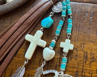 Op de afbeelding: Een zilveren sleutelhanger met een bruine leren kwast, turquoise kralen en bedels. Bedels zijn onder meer een kruis, hart, veer, hoefijzer met de tekst "Good Luck" en een schijf met de tekst "Bless this horse & rider and keep them safe."