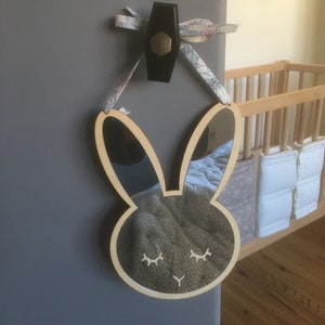 May include: A wooden bunny mirror with closed eyes, hanging on a wall with a floral ribbon. The mirror is reflecting a crib with white bedding.