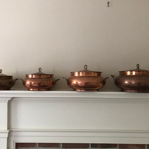 May include: Set of four copper pots with lids, arranged in size order on a white mantelpiece.