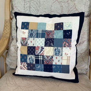 May include: A white cushion with a navy blue border and a patchwork design featuring various blue, white and red fabrics. The cushion is sitting on a beige chair with a floral pattern.