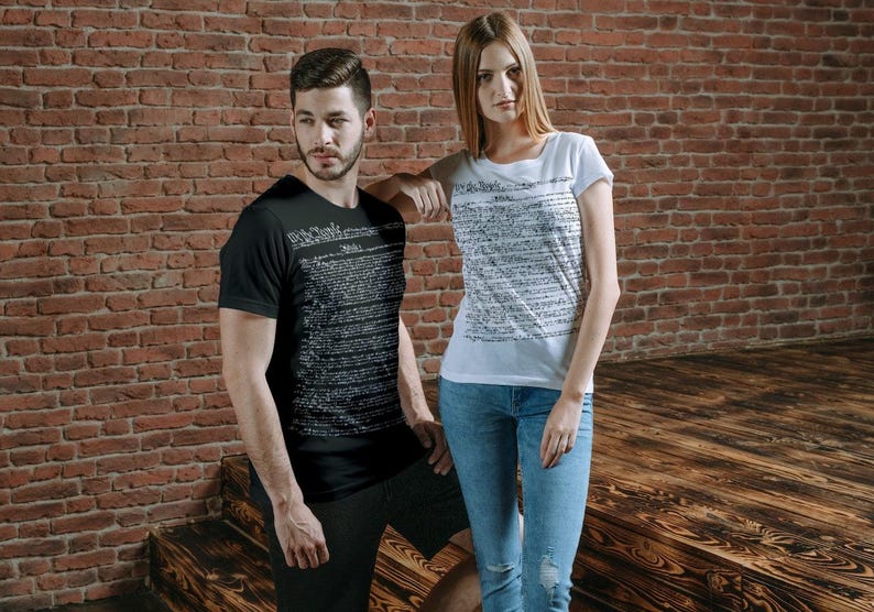 May include: Two people wearing black and white t-shirts with text printed on them. The text on the shirts is a document, possibly the US Constitution. The people are standing in front of a brick wall.