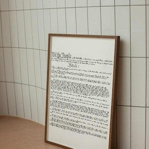 May include: A framed print of the US Constitution, featuring the text "We the People" and "Article I" in a black cursive font on a white background. The frame is brown wood.