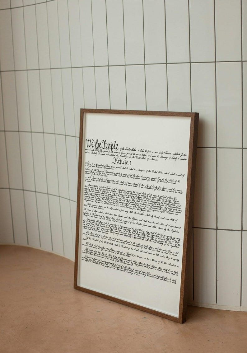 May include: A framed print of the US Constitution, featuring the text "We the People" and "Article I" in a black cursive font on a white background. The frame is brown wood.